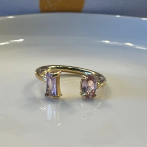 Guldring med rosa stenar - Unik guldfärgad ring med två rosa stenar i olika former – en oval och en rektangulär. Ringen har en öppen design som ger en modern och cool känsla. Perfekt för dig som vill sticka ut med ett smycke som känns både trendigt och personligt.