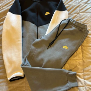 Nike tech fleece - Nike hoodie med huva och dragkedja i svart, grått och vitt. Tröjan har gul Nike-logga på bröstet, långa ärmar och är tillverkad i mjuk bomullsmix. Perfekt för chill eller träning, snygg colorblock-stil och bekväm passform.