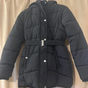 Svart pufferjacka med bälte och huva - Svart pufferjacka med quiltad design, huva och markerad midja med bälte. Jackan har långa ärmar, tryckknappar och dragkedja framtill samt praktiska fickor. Perfekt för kalla dagar när du vill hålla värmen och ändå se stilren ut. Pris kan diskuteras. 💕