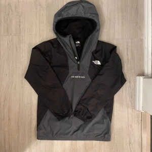 Svart och grå vindjacka The North Face - Snygg svart och grå vindjacka från The North Face med huva och halv dragkedja framtill. Jackan har logga på bröstet och ryggen, elastiska muddar och är perfekt för blåsiga dagar. Lätt och smidig att bära, passar dig som gillar streetwear och outdoor vibes.