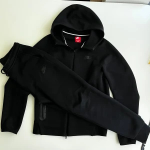 Svart Nike tech tracksuit - Svart hoodie från Nike med hel dragkedja framtill och två fickor med dragkedja. Tröjan har huva och Nike-logga på bröstet. Perfekt för dig som gillar sportig och clean stil. Materialet är mjukt och känns skönt mot huden.