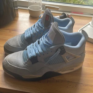 Nike Air Jordan 4 University Blue - Säljer ett par Nike Air Jordan 4 University Blue sneakers med ljusblå mocka, svarta och vita detaljer samt meshpaneler på sidorna. Skorna har klassisk snörning, platt sula och ikoniska Jordan-detaljer på plösen och hälen. Perfekta för dig som vill sticka ut med stil.