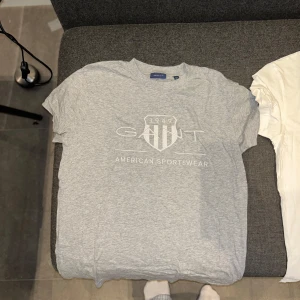 GANT t-shirt med logga, grå & vit - Två klassiska t-shirts från GANT, en i ljusgrått och en i vitt. Båda har rund hals, korta ärmar och tryckt GANT-logga med sköld och texten 'American Sportswear' framtill. Sköna och stilrena, perfekta till jeans eller shorts. 150 kr/st