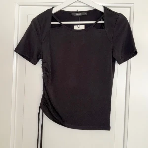 Svart topp med cut out från Bik Bok - Svart kortärmad topp från Bik Bok med cool cut out-detalj vid halsen och snörning i sidan. Toppens passform är slim och den är tillverkad i mjuk bomullsmix. Perfekt för dig som gillar trendiga detaljer och enkel stil.