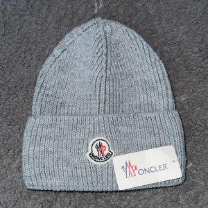 Grå stickad mössa från Moncler - Snygg grå stickad mössa från Moncler med klassisk ribbstickad design och uppvikt kant. Mössan har Moncler-logga framtill och är perfekt för att hålla värmen med stil under kyliga dagar.