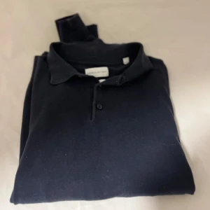 Svart pikétröja från These Glory Days - Klassisk svart pikétröja från These Glory Days i mjuk bomull. Tröjan har krage, knäppning med två knappar och en stilren look som funkar till det mesta. Perfekt för dig som gillar enkel och tidlös stil.