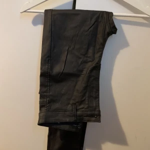 Svarta skinny jeans med glansig finish - Snygga svarta jeans med en lätt glansig finish och smal passform. Klassisk femficksmodell med bälteshällor och dragkedja. Perfekta för dig som gillar en stilren och modern look.