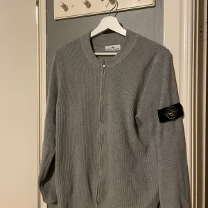 Grå stickad kofta från Stone Island - Säljer en grå stickad kofta från Stone Island med dragkedja framtill och klassisk patch på ärmen. Koftan har rund halsringning och är långärmad, perfekt för lager-på-lager-stil. Ribbade detaljer ger en clean och modern look.