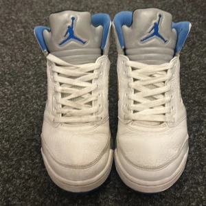 Nike Air Jordan 5 Retro vit/blå - ÄKTA* Säljer ett par feta Nike Air Jordan 5 Retro (3000kr nypris)sneakers i vitt skinn med blå detaljer och grå plös. Klassisk Jumpman-logga på plös, häl och innersula. Skorna har snörning och en chunky sula som ger en sportig vibe.  Lite missfärgning på sidan av skon så sänker därför priset