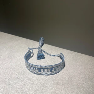 Blått armband från Dior - Snyggt blått armband från Dior med texten 'Christian Dior J'Adior' broderad i vitt. Armbandet är gjort i ett flätat tygmaterial och har justerbara tofsar för perfekt passform. Perfekt accessoar för att lyfta din outfit.