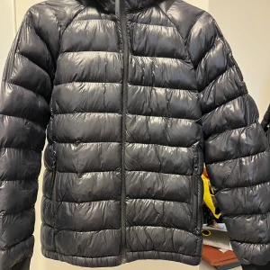 Svart dunjacka Peak Performance  - Snygg svart dunjacka från Peak Performance med horisontella sömmar, huva och dragkedja framtill. Jackan har en stilren look med logga på ärmen och är perfekt för kalla dagar. Sparsamt använd, passar både killar och tjejer! Storlek 170, köpt för 2000, pris kan diskuteras 