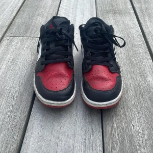 Nike Air Jordan 1 Low Bred Toe - Säljer ett par Nike Air Jordan 1 Low Bred Toe med klassisk svart, röd och vit färgkombination. Skorna har svart snörning, röd tåbox och häl, vit sidopanel samt svart Swoosh. Tillverkade i skinn med perforerad tå och ikonisk Air Jordan-logga på hälen.