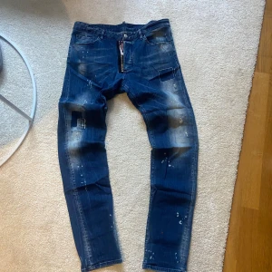 Dsquared2 jeans blå, Italiensk stl 54  - Säljer ett par Dsquared2 Cool Guy jeans i blå denim med slitningar, färgstänk och patchdetaljer. Klassisk femficksmodell med dragkedja och läderpatch bak i midjan. Snyggt tapered fit och tillverkade i Italien.