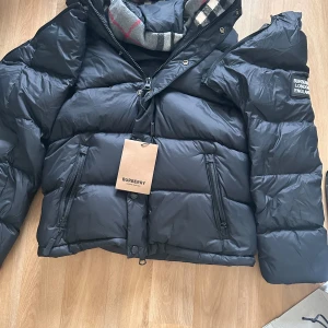 Svart pufferjacka från Burberry - Snygg svart pufferjacka från Burberry med märkets logga på ärmen. Jackan har dragkedja framtill, två stora fickor med dragkedja och hög krage. Klassisk quiltad design och en cool, oversized passform. Perfekt för kalla dagar och riktigt street.