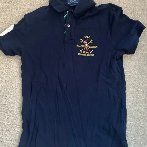 Marinblå pikétröja Ralph Lauren - Marinblå pikétröja från Ralph Lauren med broderad polospelare och RLPC-logga på bröstet. Tröjan har klassisk krage, korta ärmar och vit siffra 3 på ena ärmen. Slim fit-modell i bomull med knappar framtill och snygga detaljer.
