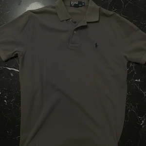 Grå pikétröja från Polo Ralph Lauren - Snygg grå pikétröja från Polo Ralph Lauren med klassisk krage och två knappar framtill. Tröjan har korta ärmar och den ikoniska broderade loggan på bröstet. Perfekt för en clean och stilren look. Sitter lite mer på den baggy sidan för M och mer tajt för L.