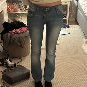 Blå bootcut jeans med ljusa detaljer - Säljer ett par blå bootcut jeans med snygga slitningar och ljusa partier framtill och baktill. Jeansen har klassisk femficksdesign, dubbla knappar i midjan och lätt utsvängda ben. Perfekta för en avslappnad och trendig look.