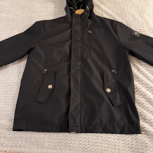 Svart vindjacka från Moose Knuckles - Svart vindjacka med huva från Moose Knuckles. Jackan har dragkedja, tryckknappar, två snedställda fickor med lock och dragkedja samt en bröstficka med dragkedja. Snygg logotyp på ärmen och justerbar huva. Perfekt för blåsiga dagar.