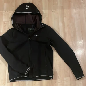 Svart Sail Racing vindjacka med huva - Svart vindjacka från Sail Racing med huva och dragkedja. Jackan har vita detaljer längs huva och ärmslut samt Sail Racing-logga på bröstet och huvan. Snygg och sportig design, perfekt för dig som gillar stilrena plagg.