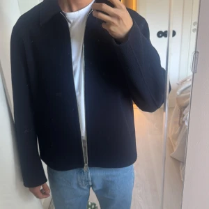 Mörkblå ulljacka med dragkedja, strl S Arket - Snygg mörkblå jacka i ull med rak passform och klassisk krage. Jackan har två snedställda fickor framtill och stängs med en silverfärgad dragkedja. Perfekt för dig som gillar stilrena och minimalistiska plagg.