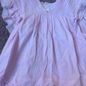 Ljusrosa klänning från Gina Tricot - Supersöt ljusrosa blus från Gina Tricot med v-ringning, volangdetaljer vid ärmarna och fina spetsinslag.  Blusen har en luftig passform. Strl 158/164 men funkar även som en XS. Använd 1 gång.