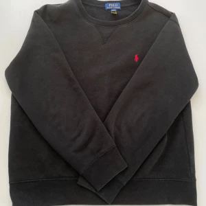 Svart sweatshirt från Polo Ralph Lauren - Svart sweatshirt från Polo Ralph Lauren med klassisk rund hals och broderad röd logga på bröstet. Tröjan har långa ärmar och är tillverkad i mjuk bomullsmix som är perfekt för kyligare dagar. Enkel och stilren design som funkar till allt.