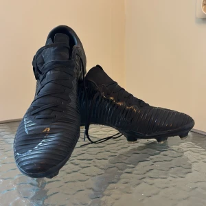 Nike Mercurial svarta fotbollsskor - Säljer ett par svarta Nike Mercurial fotbollsskor med stilren design och diskreta detaljer. Skorna har snörning, räfflad ovandel och fasta dobbar för bra grepp på planen. Perfekta för dig som vill ha en clean look på matchen. Köpta för 1200