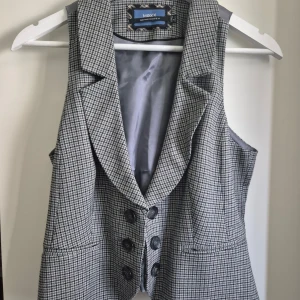 Rutig grå väst från Mexx - Snygg rutig väst från Mexx i grå och svart ton med klassisk krage och sex svarta knappar framtill. Västen har två fickor och är fodrad med ljusgrått tyg med diskreta prickar. Perfekt för att addera lite edge till din outfit. Liten i storleken som en 40:a