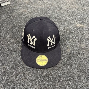 Svart New Era Yankees keps 59FIFTY - Snygg svart New Era 59FIFTY keps med New York Yankees loggor broderade runt hela kepsen och årtal under varje logga. Platt skärm och klassisk passform. Materialet är 100% ull och kepsen har en gul etikett på skärmen. Perfekt för dig som gillar streetwear och baseball.