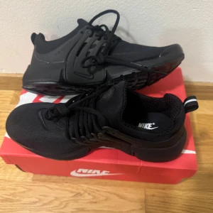 Svarta Nike Presto sneakers - Nike Presto sneakers i helsvart färg med snygg, strömlinjeformad design. Ovandel i mesh och syntet, med rund tå och platt sula. Skorna har snörning och en diskret Nike-logga på sidan. Perfekt för dig som gillar stilrena och sportiga sneakers. Box ingår.