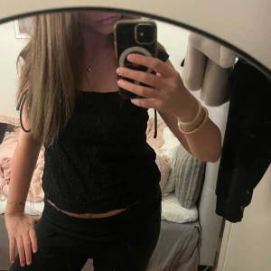 Svart smockad linnetopp Gina Tricot - Svart linnetopp från Gina Tricot i smockat tyg med tjocka axelband och snören som man kan knyta med på axlarna. Toppen är croppad och har en enkel, stilren look som passar perfekt till jeans eller kjol. Mjuk och lätt att bära, perfekt för varma dagar.