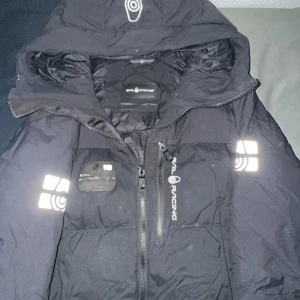 Svart Sail Racing dunjacka med huva - Svart dunjacka från Sail Racing med huva och reflekterande detaljer på ärmarna. Jackan har tvåvägsdragkedja framtill, logga på bröstet och patch med 'Antarctica' på vänster sida. Perfekt för kalla dagar och riktigt snygg streetstil.