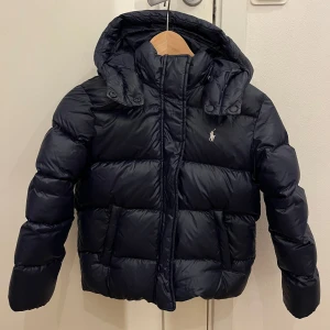 Mörkblå dunjacka från Polo Ralph Lauren barnstorlek 8-10 år - Snygg mörkblå dunjacka från Polo Ralph Lauren med broderad logga på bröstet för barn. Jackan har huva, dragkedja och två fickor framtill. Perfekt för kalla dagar och riktigt skön att bära. Klassisk pufferstil med quiltade sektioner. Storlek 8-10 år. Väldigt fint skick.