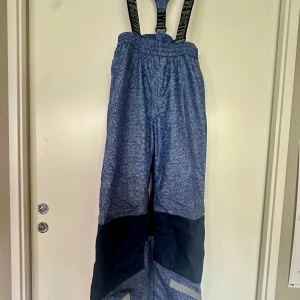 Blå skalbyxor från Mywear Active - Blå skalbyxor från Mywear Active med svarta detaljer på benen och justerbara hängslen. Byxorna har elastisk midja och reflekterande detalj nedtill. Perfekta för kalla dagar och utomhusaktiviteter.