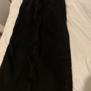 Svart lång jeanskjol med slits - Säljer en svart lång jeanskjol med klassisk rak passform och slits framtill. Kjolen har fickor bak och är tillverkad i ett robust denimtyg. Perfekt för dig som gillar en stilren och tidlös look.