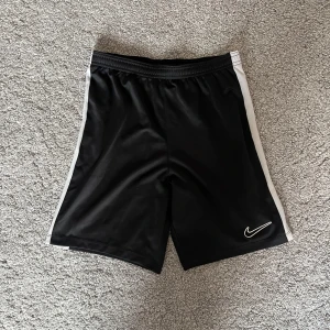 Svarta sportshorts från Nike - Svarta sportiga shorts från Nike med vita sidor och elastisk midja. Tillverkade i ett glansigt, lätt polyestermaterial som andas. Klassisk Nike-logga broderad på ena benet. Perfekta för träning eller chill dagar.