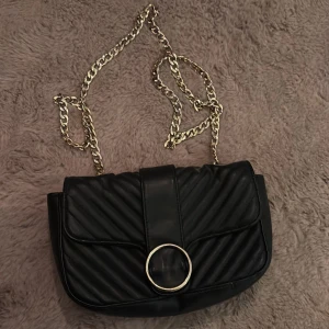 Svart quiltad axelväska med guldkedja - Snygg svart axelväska i quiltat skinn med guldfärgad kedja och rund metallring framtill. Väskan har ett stilrent och trendigt utseende, perfekt för att lyfta vilken outfit som helst. Praktisk storlek för det viktigaste och enkel att bära över axeln.