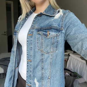 Blå jeansjacka från Stradivarius - Snygg blå jeansjacka från Stradivarius i storlek S. Jackan har slitna detaljer vid axlar och på flera ställen, klassisk krage, två bröstfickor med knappar och knappstängning framtill. Perfekt för dig som gillar en chill och trendig look.