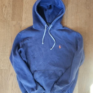 Blå hoodie från Polo Ralph Lauren - Säljer en blå hoodie från Polo Ralph Lauren med vit snörning och klassisk orange logga broderad på bröstet. Tröjan har huva, känguruficka och är tillverkad i mjuk bomull. Perfekt för chill dagar eller när du vill ha en clean och stilren look.