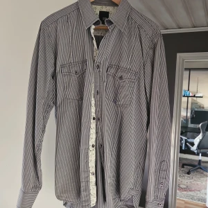 Randig skjorta från Jack & Jones L - Snygg randig skjorta från Jack & Jones i storlek L. Skjortan är blåvit med smala ränder, har två bröstfickor med knappar och klassisk krage. Tillverkad i bomull och har långa ärmar. Perfekt för en stilren look.