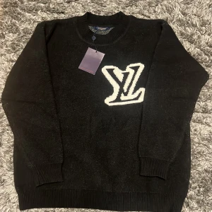 Svart stickad tröja från Louis Vuitton  - Svart stickad tröja från Louis Vuitton med stort vitt LV-logo framtill. Tröjan har rund halsringning, ribbade muddar och är långärmad. Perfekt för dig som vill ha en lyxig och stilren look med streetkänsla.