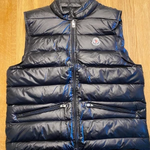 Moncler Gui väst Svart - Svart dunväst från Moncler med glansig finish och horisontella quiltade paneler. Västen har två snedställda fickor med dragkedja och en hög krage. Moncler-logga på bröstet och hel dragkedja framtill. Perfekt för lager-på-lager under kyliga dagar.                                                         Pris går att diskutera!