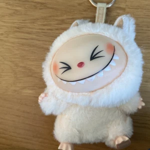 Labubu Pop Mart - Supergullig liten labubu från Pop Mart formad som ett monster med stort leende. Pälsig och mjuk i vitt syntetmaterial, med öron och broderade detaljer. Perfekt som accessoar eller nyckelring till väskan. Hängs enkelt med metallring.