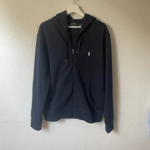 Svart hoodie från Polo Ralph Lauren - Klassisk svart hoodie från Polo Ralph Lauren med vit broderad logga på bröstet. Tröjan har dragkedja, huva med snörning och två fickor framtill. Perfekt för en chill och stilren look.