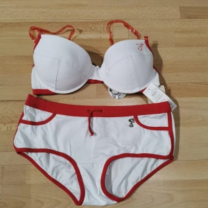 Snoopy bh och hipster vit och röd - Superfin set från Snoopy med vit bas och röda detaljer. Bh  har en vadderad överdel med transparenta axelband och en söt Snoopy-charm framtill. Underdelen har röda kanter, snörning i midjan och ett  Snoopy-motiv bak. Materialet är mjuk bomull med lite stretch.