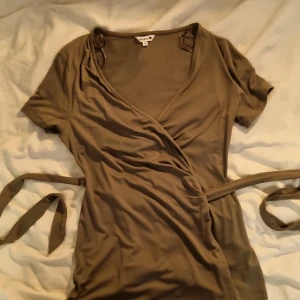 Olivgrön omlottklänning från Indiska XS - Supersnygg olivgrön omlottklänning från Indiska i mjukt material. Klänningen har korta ärmar, v-ringning och knytband i midjan för en smickrande passform. Perfekt för dig som gillar enkel och stilren design.