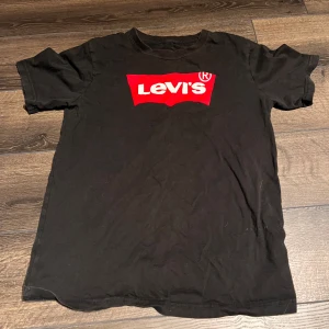Svart Levi's t-shirt med röd logga - Svart t-shirt från Levi's med klassisk röd logga framtill. T-shirten har rund halsringning och korta ärmar. Tillverkad i mjuk bomull för en skön känsla. Perfekt för en avslappnad och cool stil.