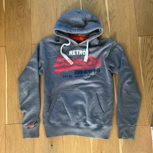 Grå Superdry hoodie med tryck - Grå hoodie från Superdry med rött och vitt retro-tryck på bröstet och japanska tecken. Tröjan har huva med vita snören, känguruficka och ribbade muddar. Perfekt för dig som gillar streetstyle och vill ha en snygg och bekväm look slim/ straight fit