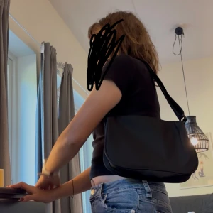 Svart axelväska i syntetmaterial - Snygg svart axelväska med justerbart band och enkel, stilren design. Väskan är tillverkad i slätt syntetmaterial och har en mjuk, rundad form. Perfekt storlek för att bära med sig det viktigaste och passar till många olika outfits.