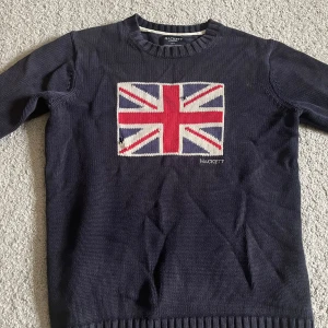 Mörkblå stickad tröja Hackett London - Mörkblå stickad tröja från Hackett London med Union Jack-flagga i rött, vitt och blått på bröstet. Perfekt för dig som gillar brittisk stil och vill sticka ut med en klassisk look.
